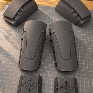 May include: A set of six dark gray foam armor pieces including two curved upper arm guards, two forearm guards with rivet details, and two smaller rectangular wrist guards featuring a geometric embossed design.