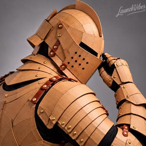 May include: A suit of armour constructed from tan corrugated cardboard, featuring a helmet with a visor, segmented shoulder plates, and articulated arm guards. The pieces are joined by brown leather straps and metallic gold rivets.