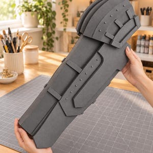 Puede incluir: Un protector de brazo de espuma gris oscuro con una hombrera superpuesta, una sección larga de antebrazo con paneles rectangulares elevados y una tira central con pequeños remaches circulares. El texto LaunchVibes! aparece abajo.