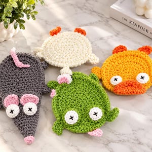 Peut inclure: Quatre dessous de verre en crochet faits main en forme d'animaux de dessin animé. Il y a une souris grise, une créature crème, une figure orange vif ressemblant à un canard et une grenouille verte, avec des yeux blancs et des croix noires.