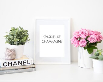 May include: A white framed print with the text "Sparkle Like Champagne" on a white table with a potted plant, a candle, and a bouquet of pink roses in a white vase.