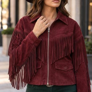 May include: A deep burgundy suede jacket featuring long fringe detailing across the chest, back, and sleeves. The jacket includes a silver-toned front zipper closure, a classic collar, and two front flap pockets with button closures at the waist.