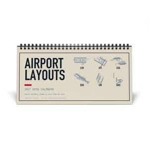 Può includere: Un calendario da scrivania 2027 con rilegatura a spirale sulla parte superiore. La copertina è beige con testo blu scuro che recita AIRPORT LAYOUTS e 2027 DESK CALENDAR. Presenta diagrammi tecnici blu di sei aeroporti etichettati LHR, JFK, CDG, DXB, SIN e AMS.