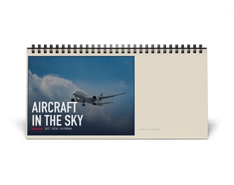 May include: A 2027 desk calendar with a black spiral binding. The cover features a photograph of an airplane flying through a cloudy sky, with the white text AIRCRAFT IN THE SKY and 2027 DESK CALENDAR printed below the image on a dark background.