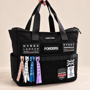 May include: A black tote bag featuring multiple patches including Hyrox London, Hyrox Birmingham, Forders, and Carly Club. The bag has black handles, side pockets, and four hanging ribbons in pink, white, blue, and purple with Hyrox branding.