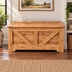 Puede incluir: Un baúl de almacenamiento rectangular de madera con un acabado marrón claro y veta de madera visible. El panel frontal presenta un soporte vertical central flanqueado por dos refuerzos diagonales, creando un patrón. El baúl tiene una superficie superior plana.