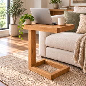 Puede incluir: Una mesa auxiliar en forma de C hecha de madera de tono claro con una superficie rectangular y una base resistente. La mesa sostiene un ordenador portátil plateado y una taza de cerámica blanca. El diseño presenta líneas limpias y veteado.