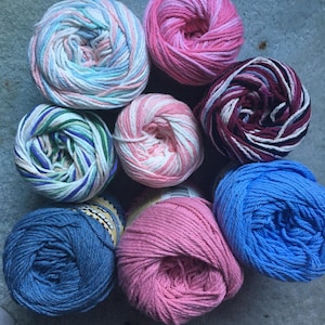 May include: Eight skeins of yarn in various colors and patterns. The collection includes solid blue, solid pink, variegated pink, variegated blue and white, variegated green and purple, and a multi-colored skein with dark red, white, and black.