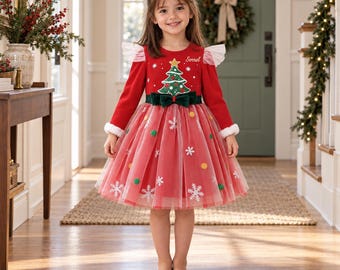 May include: A red long-sleeved dress featuring a Christmas tree graphic with the name Sorrel, a dark green velvet waist bow, and a layered tulle skirt adorned with white snowflake appliques and small colorful pom-pom ornaments.