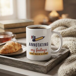 Puede incluir: Una taza de cer&aacute;mica blanca con un gr&aacute;fico de un l&aacute;piz amarillo y pesta&ntilde;as de colores. El texto "ANNOTATING my feelings ONE TAB AT A TIME" aparece en azul oscuro, naranja y amarillo. La taza tiene un asa est&aacute;ndar y un acabado liso.
