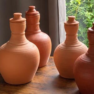 May include: Four terracotta clay vessels with rounded bodies and narrow necks, each topped with a small matching lid. The vessels feature a smooth, matte finish in a warm, earthy orange-brown tone, arranged in a cluster on a wooden surface.