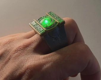 May include: A metallic ring featuring a dark grey band and a rectangular top section with a textured gold-toned border. The center of the ring contains a circular, translucent green element that emits a bright, glowing light from within.