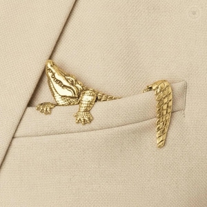 May include: A gold-toned metal crocodile brooch pinned to a beige blazer pocket. The brooch is designed in two separate pieces, with the head and front legs emerging from the pocket and the textured tail section attached separately.