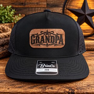 Könnte beinhalten: Eine schwarze Baseballkappe mit einem rechteckigen Korkaufnäher, der das Wort GRANDPA in schwarzer Schrift und ein Sternsymbol zeigt. Die Kappe hat einen flachen Schirm und einen schwarzen Aufkleber mit dem Text BrimCo EST. 2025 QUALITY 200 HEADWEAR.