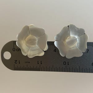 May include: Two white iridescent mother-of-pearl flower-shaped earrings resting on a metal ruler. The ruler shows each flower measures approximately 1.5 inches in diameter. The petals have a smooth, glossy finish and a central circular core.