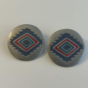 May include: A pair of round, silver-toned metal earrings featuring a central geometric diamond pattern. The design includes concentric layers of dark blue, red, and teal enamel set within the brushed metal surface of each circular earring.