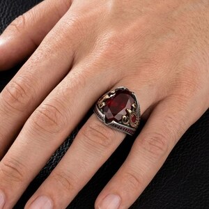 May include: A silver ring featuring a large, oval-cut dark red gemstone set in a decorative metal band with intricate engravings and small red accents on the sides, worn on the ring finger of a hand.