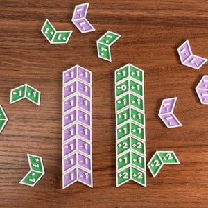 May include: A collection of chevron-shaped game tokens in purple and green. Two vertical columns are formed by stacked tokens, while individual tokens are scattered around. Each token features white numerical text including -1, +0, +1, and +2.