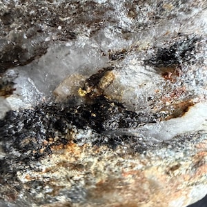 May include: A raw mineral specimen featuring a translucent white quartz matrix with prominent veins of dark, textured inclusions and small, metallic gold-colored flecks embedded within the crystalline structure of the rock surface.