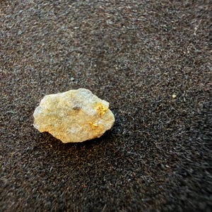 May include: A small, irregular piece of light gray, textured rock featuring visible flecks of metallic gold embedded within its surface. The stone has a rough, uneven appearance with a matte finish and distinct mineral inclusions throughout its structure.