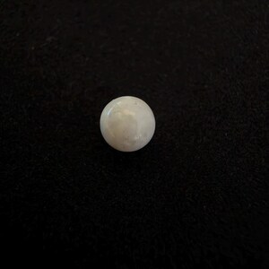 May include: A single, spherical, off-white pearl with a smooth, lustrous surface. The object is round and exhibits a subtle, pearlescent sheen, reflecting light across its curved exterior. It is presented as a solitary, small, circular item.