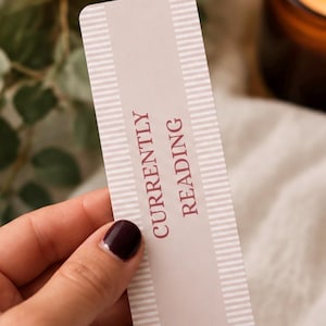 May include: A rectangular bookmark featuring a light beige background with white horizontal stripes along the top and bottom edges. The text "CURRENTLY READING" is printed in a dark maroon, sans-serif font vertically along the center.
