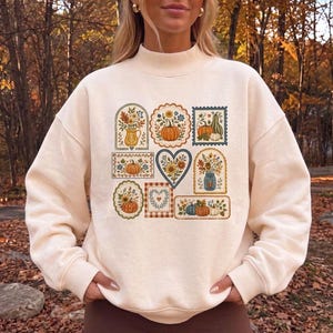 Peut inclure: Un sweat-shirt crème à col montant orné d'un imprimé graphique central composé de diverses illustrations encadrées, incluant des citrouilles, des fleurs d'automne et du feuillage dans des tons orange, bleu, jaune et vert.