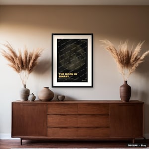 Pode incluir: Uma impressão de arte retangular emoldurada com um fundo escuro e um gráfico de lua dourada texturizada. O texto THE MOON IN BINARY está impresso em amarelo no centro inferior. A impressão está numa moldura preta simples com paspatur branco.