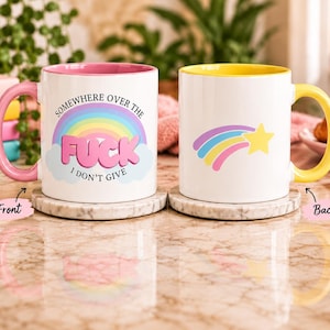 May include: Two white ceramic mugs with coloured interiors and handles. The left mug has a pink handle and a rainbow graphic with the text 'SOMEWHERE OVER THE FUCK I DON'T GIVE'. The right mug has a yellow handle and a rainbow shooting star graphic.