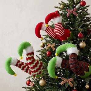 May include: Three pairs of plush elf legs protruding from a Christmas tree. One pair features red and white horizontal stripes, another has a red, white, and green chevron pattern, and the third has a dark red and brown knit pattern.