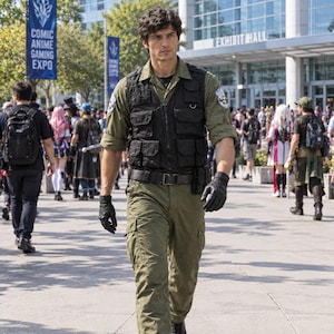 May include: A person wearing an olive green button-down shirt with rolled sleeves, a black tactical vest with multiple pockets, matching olive green cargo pants, a black belt, and black gloves. The vest features a circular patch on the left shoulder.