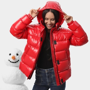 Puede incluir: Una chaqueta acolchada de color rojo brillante con acabado de alto brillo, capucha y cierre de cremallera frontal con detalles en negro. La prenda cuenta con mangas largas con puños elásticos y un cuello alto, ofreciendo un aspecto reflectante.
