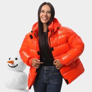Puede incluir: Una chaqueta acolchada de color naranja brillante y acabado de alto brillo, con capucha, mangas largas con puños elásticos y cierre frontal de cremallera con tiras de velcro negras. La prenda presenta un diseño acolchado.