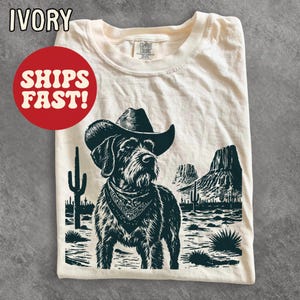 May include: An ivory t-shirt featuring a dark graphic print of a dog wearing a cowboy hat and a bandana. The background illustration depicts a desert landscape with cacti and rock formations. The shirt has a crew neckline and a Comfort Colors label.