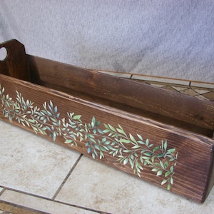 May include: A long, rectangular dark brown wooden box with a hand-painted design of green and blue leaves along the front side. The box features two raised end panels, each with a circular cutout handle, and a deep, open-top storage compartment.