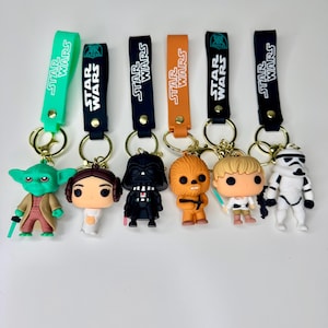 May include: Six Star Wars character keychains featuring Yoda, Princess Leia, Darth Vader, Chewbacca, Luke Skywalker, and a Stormtrooper. Each figure is attached to a coloured silicone strap with the Star Wars logo and a gold-toned metal keyring.