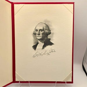 May include: A red-bound book or folder featuring an off-white interior page with cream-colored corner tabs. The page displays a black and white portrait of George Washington, accompanied by his signature, G. Washington, written in cursive script below.