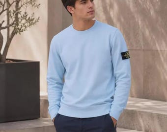 Op de afbeelding: Een lichtblauw sweatshirt met lange mouwen, een ronde hals en een zwart, geel en groen kompasvormig embleem op de linkermouw. Het kledingstuk wordt gecombineerd met een donkerblauwe broek voor een eenvoudige en strakke uitstraling.