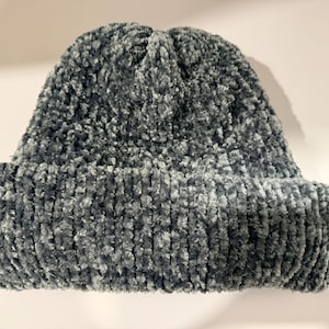 Puede incluir: Un gorro tejido con hilo de felpa texturizado en una paleta de colores gris oscuro y azul pizarra. El gorro presenta un borde grueso doblado y una construcción de punto denso y difuso que crea una apariencia suave y táctil.