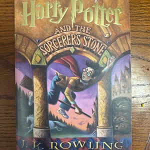 May include: A paperback book titled Harry Potter and the Sorcerer's Stone by J.K. Rowling. The cover features an illustration of a young person with glasses flying on a broomstick through a stone archway, with a unicorn and castle visible.