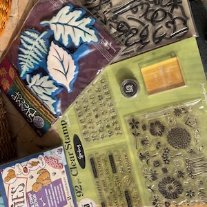 May include: A collection of craft supplies including a Stamp Decor leaf stamp set, a 92-piece clear stamp set with floral and alphabet designs, a small Ink Dot ink pad, a clear mounting block, and a Doggies rubber stamp collection box.