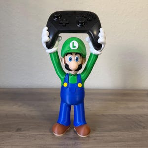 May include: A Luigi figurine with a green hat featuring a white L, a green shirt, blue overalls with yellow buttons, and brown shoes. The figure has white gloved hands raised above its head, holding a black wireless video game controller.