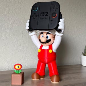May include: A Mario figure with a white hat, white shirt, red overalls, and brown shoes holding a black Nintendo Switch console above its head. Next to the figure is a small brown cube base topped with a red and yellow flower icon.