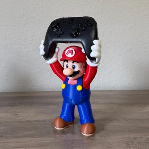 May include: A 3D printed figure of the character Mario wearing a red hat with an M logo, a red shirt, blue overalls with yellow buttons, and brown shoes. The figure holds a black wireless video game controller above its head with white gloved hands.