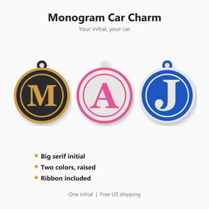 May include: Three circular monogram car charms featuring a large serif initial in the center. The first is black with a gold M, the second is white with a pink A, and the third is white with a blue J. Each charm has a small loop at the top.