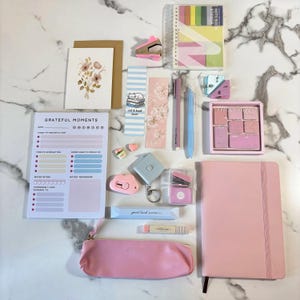 May include: A collection of stationery items including a Grateful Moments notepad, a pink pencil case, a pink notebook, various sticky notes, bookmarks, pens, a small stapler, a tape measure, and a floral greeting card arranged on a surface.