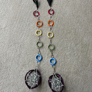 May include: Two vertical hanging ornaments featuring a series of small fabric-wrapped rings in red, orange, yellow, green, and blue, connected by silver metal jump rings. Each ends with a larger purple hoop containing a silver webbed design.