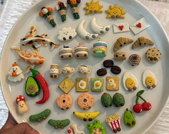 May include: A collection of small, colorful clay charms arranged on a white plate, featuring various shapes including koi fish, suns, moons, food items like toast, eggs, fruits, vegetables, and sweets, all with simple, hand-painted smiling faces.
