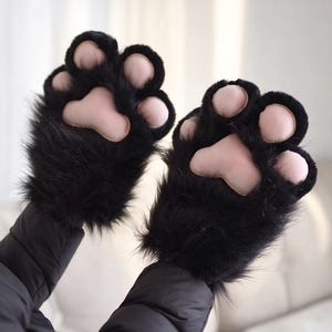 May include: A pair of black faux fur fingerless gloves designed to resemble animal paws. Each glove features soft, light pink padded paw pads and toe beans, with long black synthetic fur covering the back of the hand and wrist area.