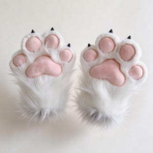 May include: A pair of white faux fur gloves shaped like animal paws. Each glove features four soft, light pink padded paw pads and small, pointed black claws at the tips of each finger, mimicking the appearance of a feline or canine paw.